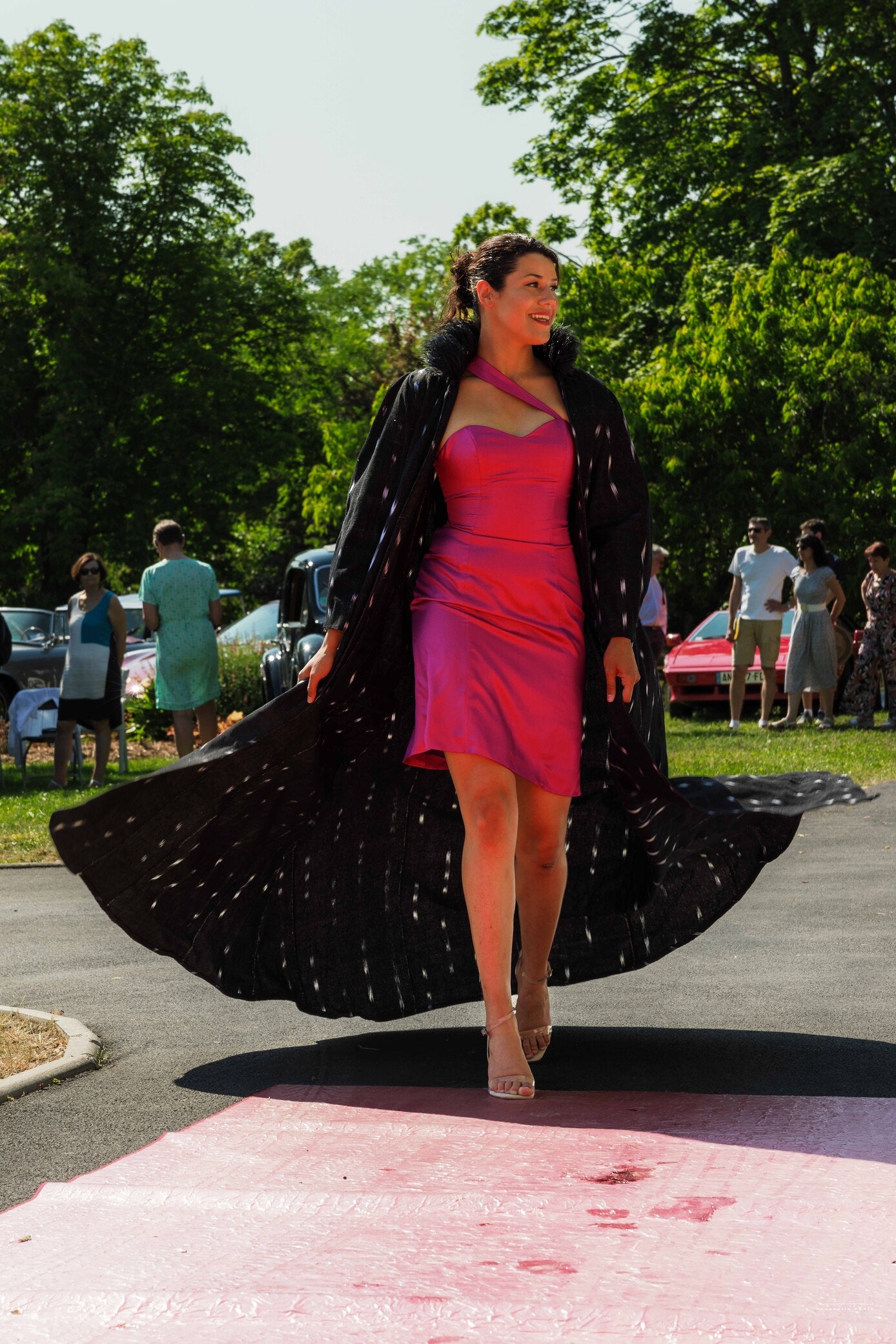 Ensemble Opéra Ousman O. avec robe courte en soie fuchsia à bretelle asymétrique et cape-manteau noire à motifs clairs, col Mao avec plumes d’autruche noire, présenté lors d’un défilé chez Champagne Gosset.
